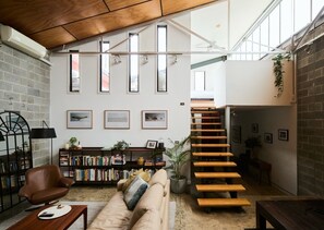 A spacious loft with a high ceiling, a staircase, a cozy seating area with a sofa and armchair, and a bookshelf filled with books. - Bottega Loft Industrial Charm with Artistic Flair (Brunswick)