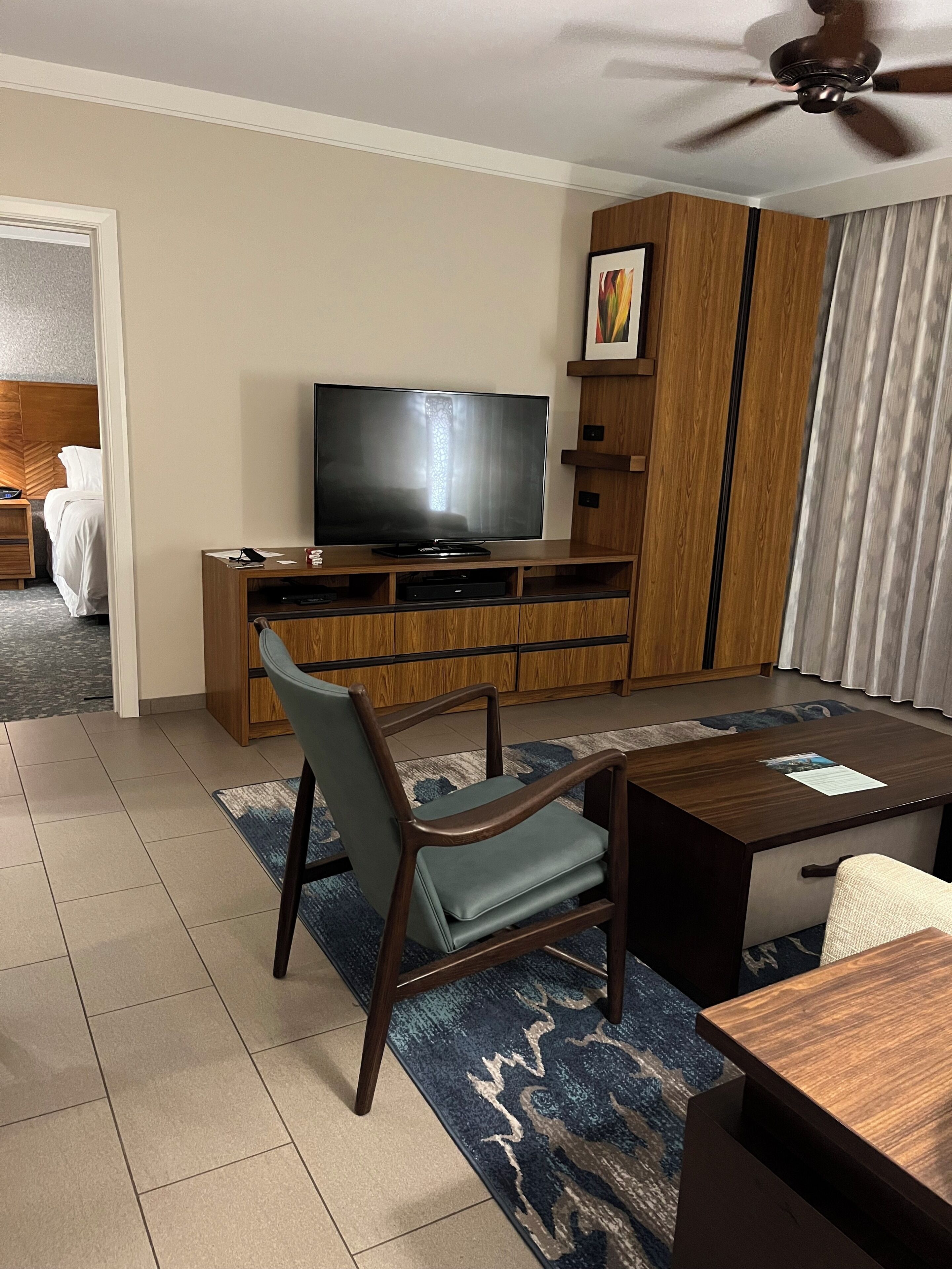 Quarto de hotel com televisão de tela plana em um suporte de madeira, mesa de jantar com cadeiras, ventoinha no teto e vista para o corredor.