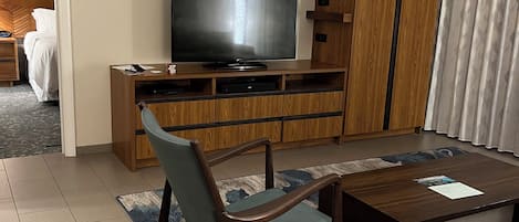 Habitación de hotel con televisor de pantalla plana sobre un soporte de madera, una mesa de comedor con sillas, un ventilador de techo y vistas al pasillo.