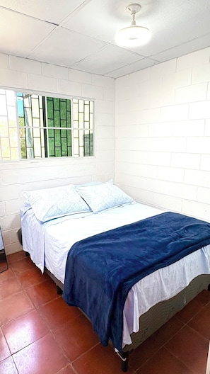 Ein Zimmer mit einem Bett, weißen Backsteinwänden, einem Fenster mit grün getöntem Glas und einem Deckenventilator.
