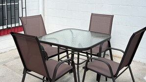 Eine Terrasse im Freien mit einem runden Glastisch und vier Stühlen, einer weißen Backsteinmauer und einem Fenster mit schwarzen Streben.