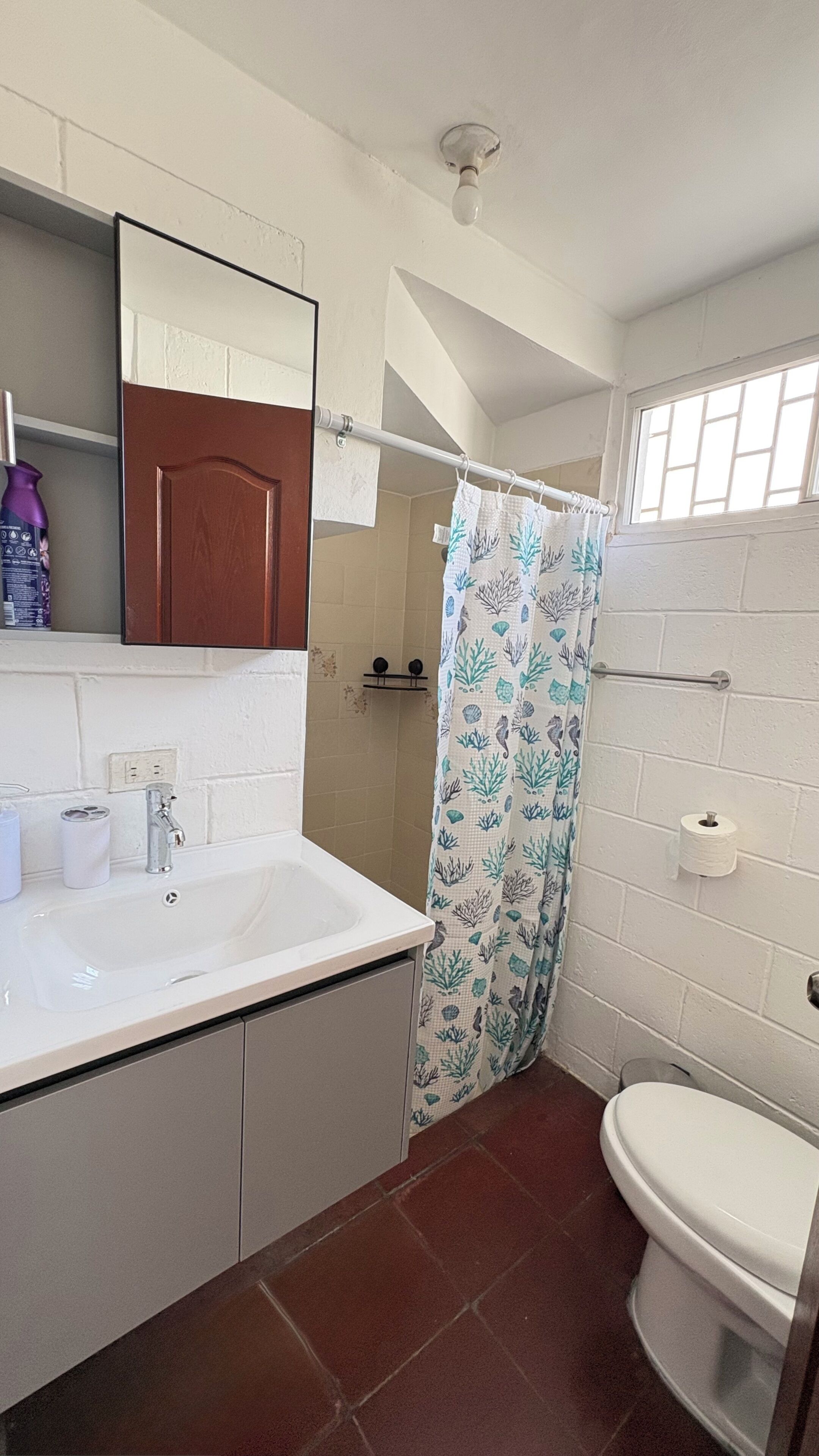 Un baño con lavamanos blanco, inodoro, cortina de ducha con motivos marinos, espejo y ventana.