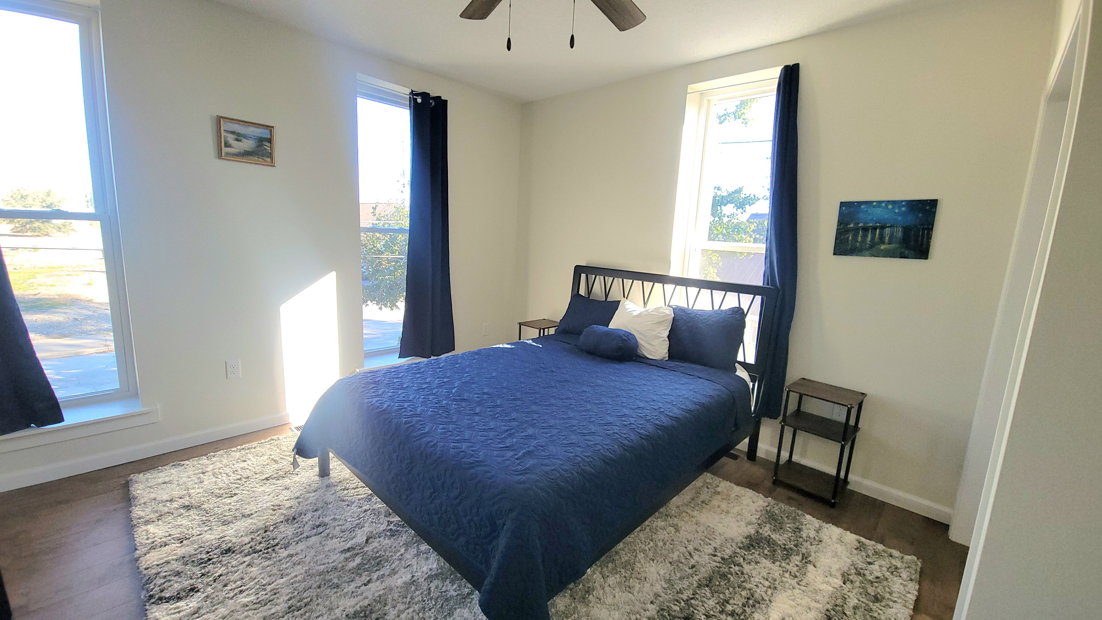 Un dormitorio con cama, mesita de noche, ventana con cortinas y ventilador de techo.