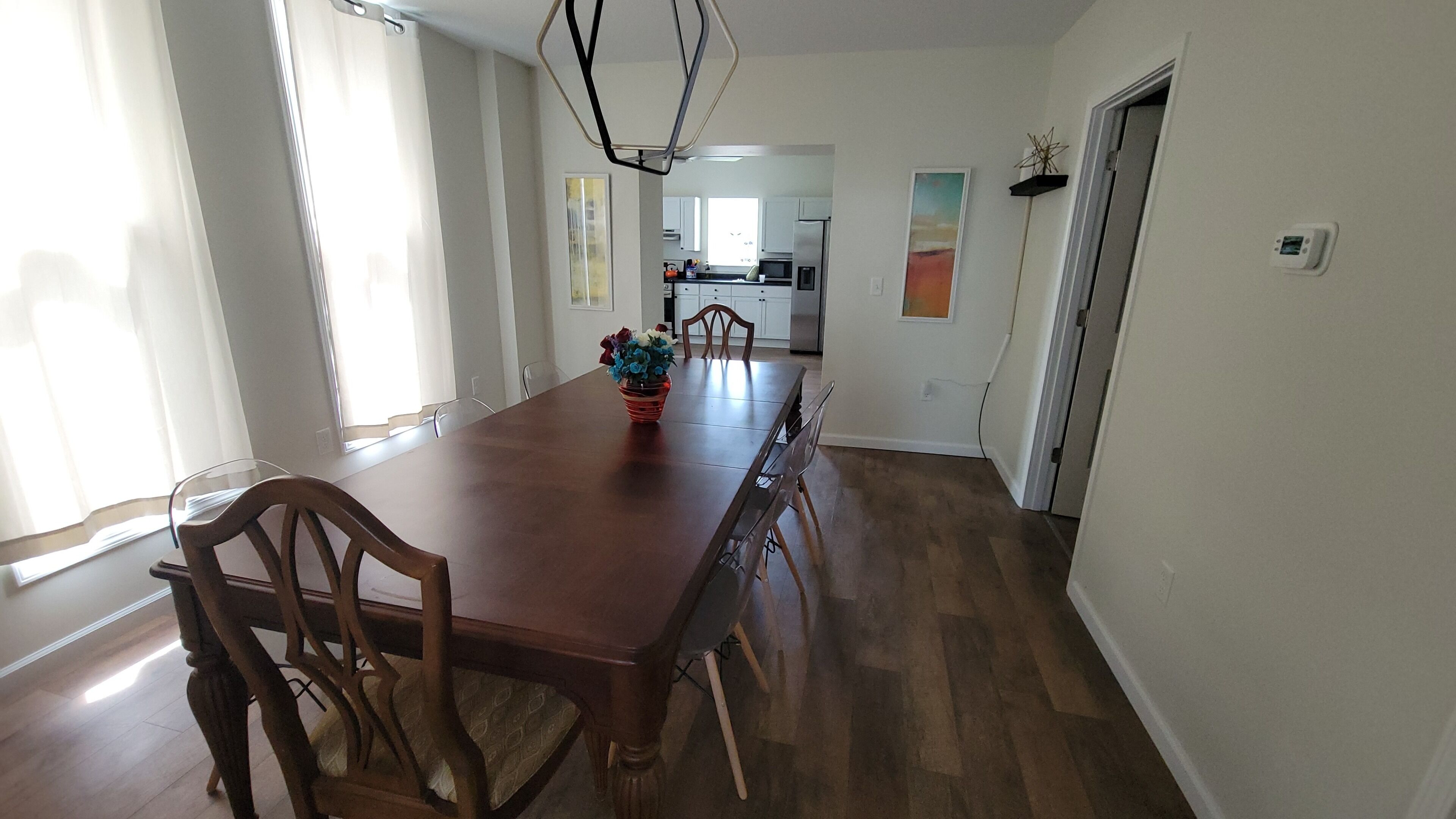 A dining room with a wooden table and chairs, a vase with flowers, and a view into the kitchen.
