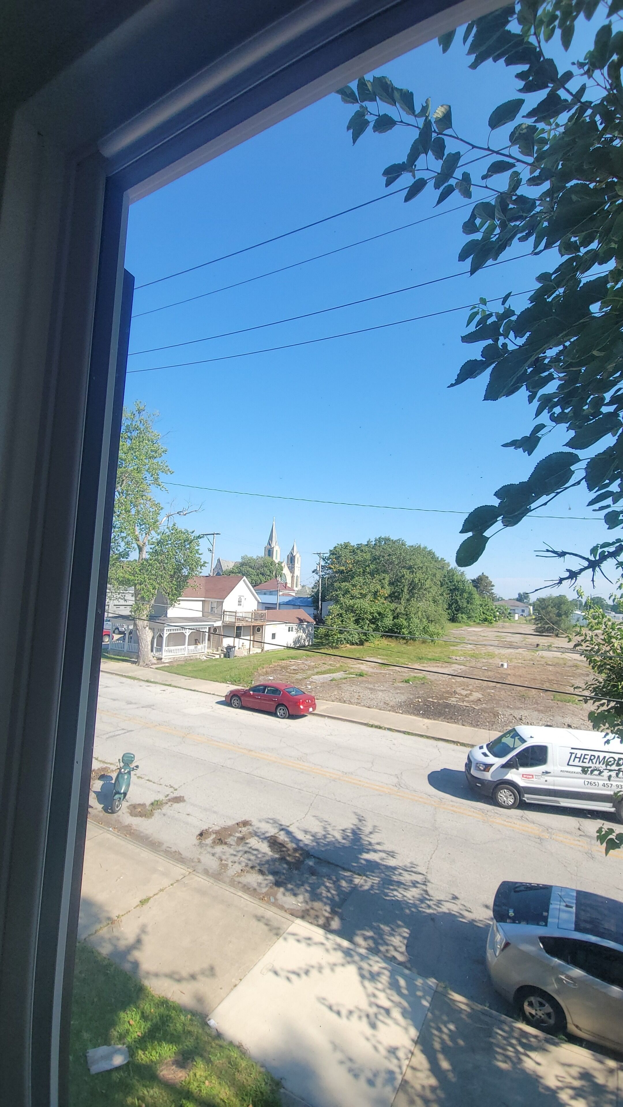 Una vista attraverso una finestra mostra una strada con un'auto rossa parcheggiata sul lato, un furgone bianco e un edificio con un campanile in secondo piano.