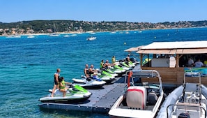 Ein Bereich zur Jet-Ski-Verleih mit mehreren Jet-Skis, die an einem Steg liegen, einer kleinen Holzkonstruktion und Blick auf das Meer und die Küste in der Ferne.