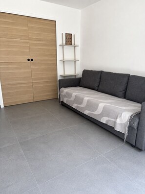 Ein Zimmer mit grauem Sofa, einem Holzschrank und einer weißen Wand mit Regal.