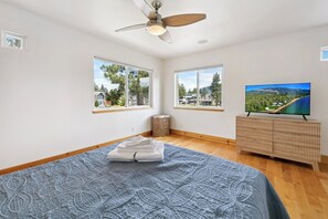 Ein Schlafzimmer mit einem Bett, einem Fernseher und zwei Fenstern mit Blick auf Häuser und Bäume.