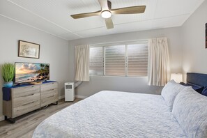 A bedroom with a bed, a ceiling fan, a window with curtains, a TV on a dresser, and a plant. - NEW- Renovated Ocean view- Across from the beach- Fantastic location (Kihei)