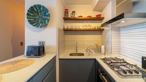 Uma cozinha compacta com fogão a gás, pia e torradeira. Há prateleiras acima da pia com diversos utensílios de cozinha.