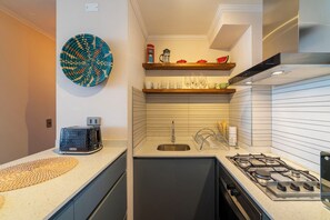 A compact kitchen with a gas stove, a sink, and a toaster. There are shelves above the sink holding various kitchen items. - Americo Vespucio Apartments By Andes STR (Las Condes)