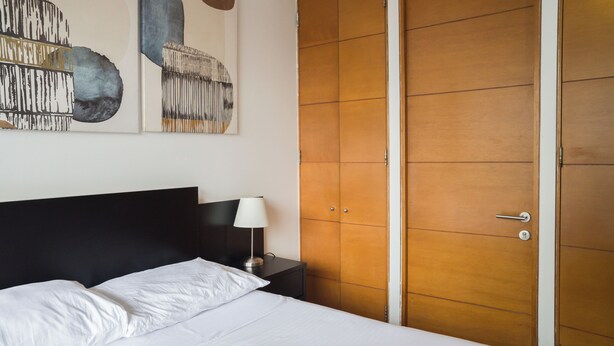 Quarto de hotel com cama, mesa de cabeceira, luminária e guarda-roupa.
