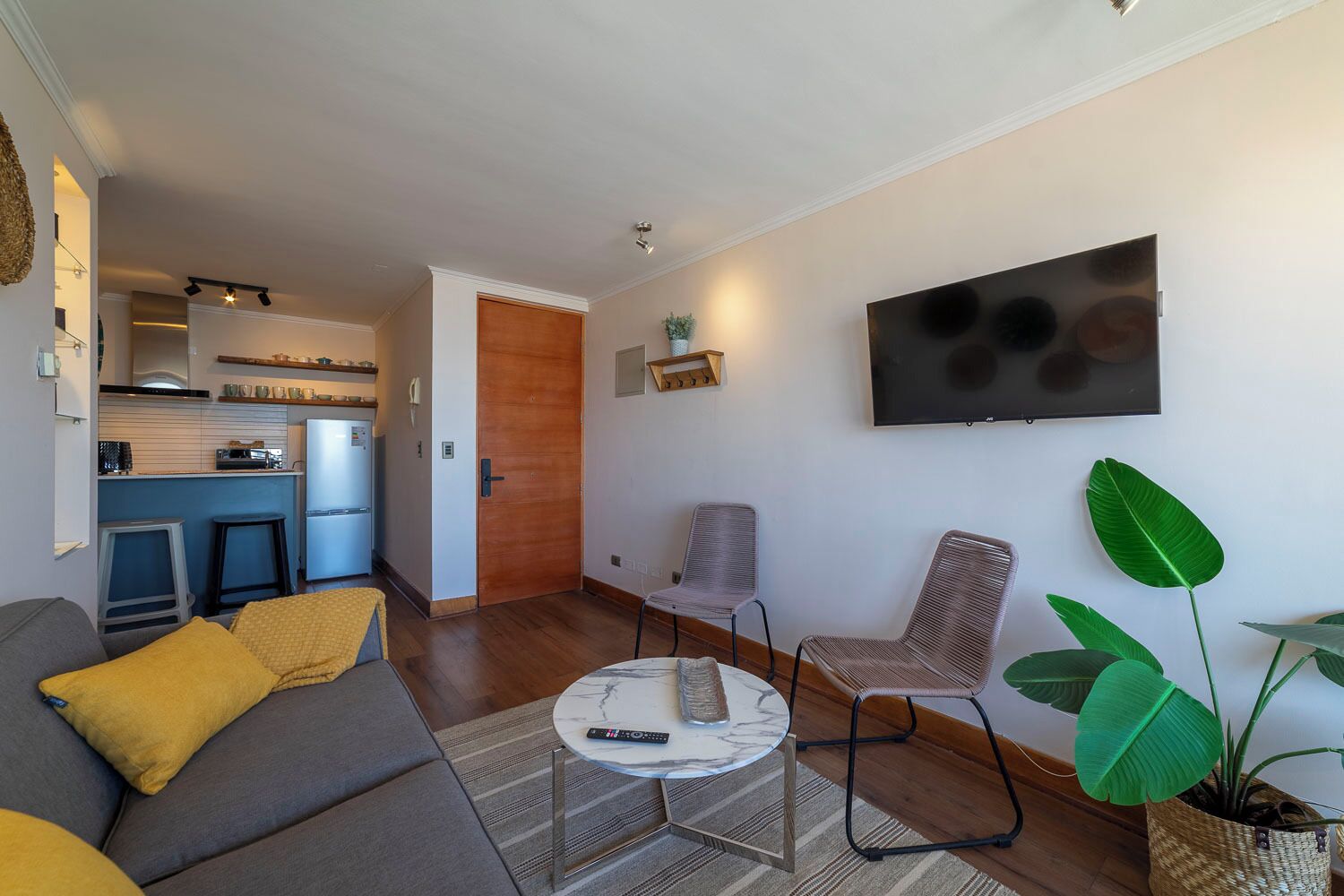 Ein Wohnzimmer mit einer Couch, einem Couchtisch und zwei Sesseln. An der Wand ist ein Fernseher angebracht und in der Ecke steht eine Pflanze.