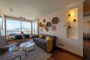 Ein modernes Wohnzimmer mit einer Couch, Sesseln, einem Couchtisch und einem Essbereich mit Tisch und Stühlen. An den Wänden befinden sich Wandgestaltungen und ein Regal mit Gegenständen.