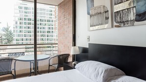 Quarto de hotel com cama, mesa pequena, duas cadeiras, varanda e vista para o horizonte urbano, além de obras de arte na parede.