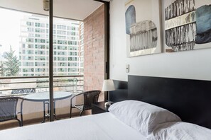A hotel room with a bed, a small table, two chairs, a balcony with a view of a cityscape, and artwork on the wall. - Americo Vespucio Apartments By Andes STR (Las Condes)