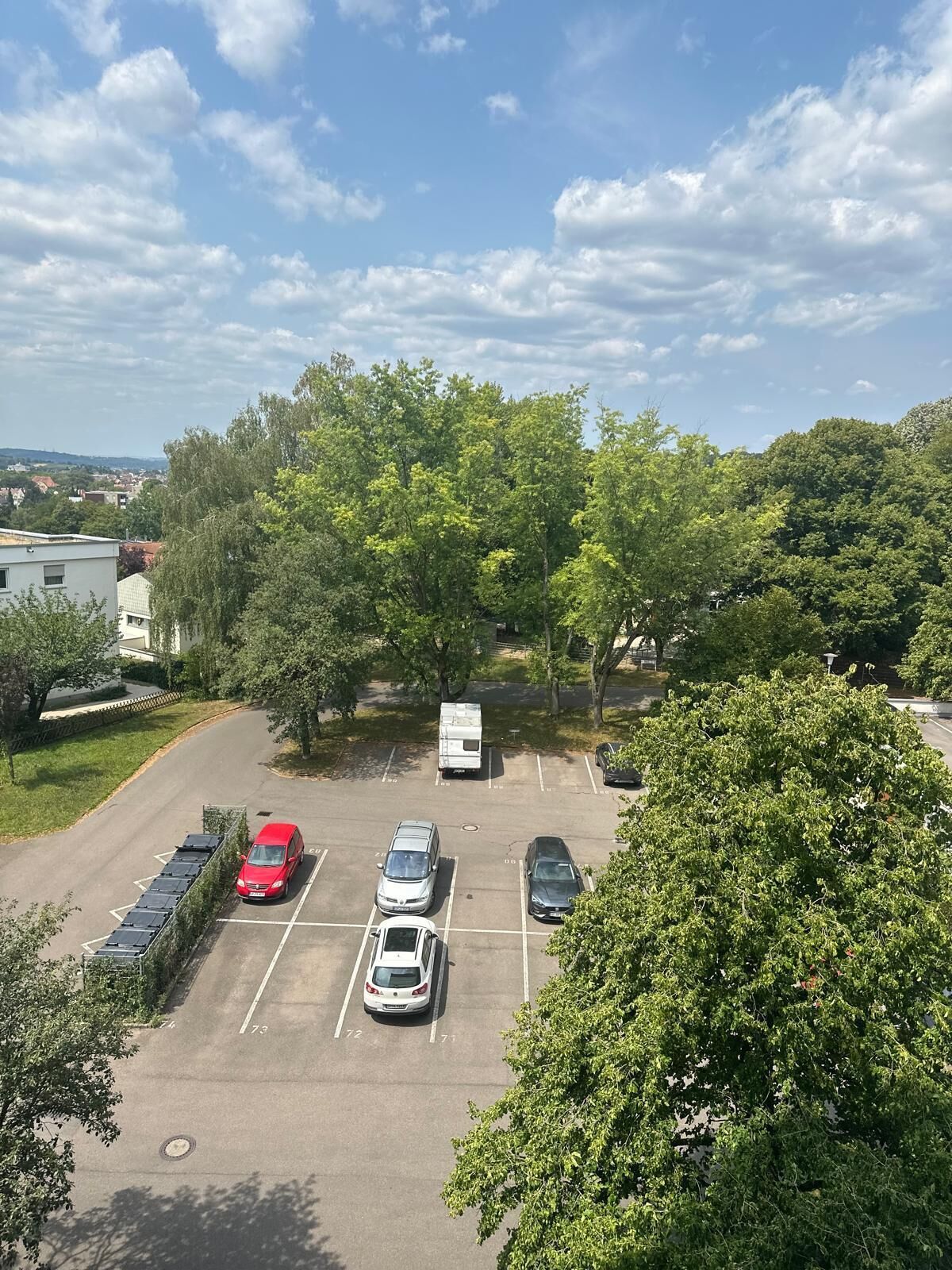 Ein Parkplatz mit mehreren Autos und einem weißen Lieferwagen, umgeben von Bäumen und einem Gebäude im Hintergrund.