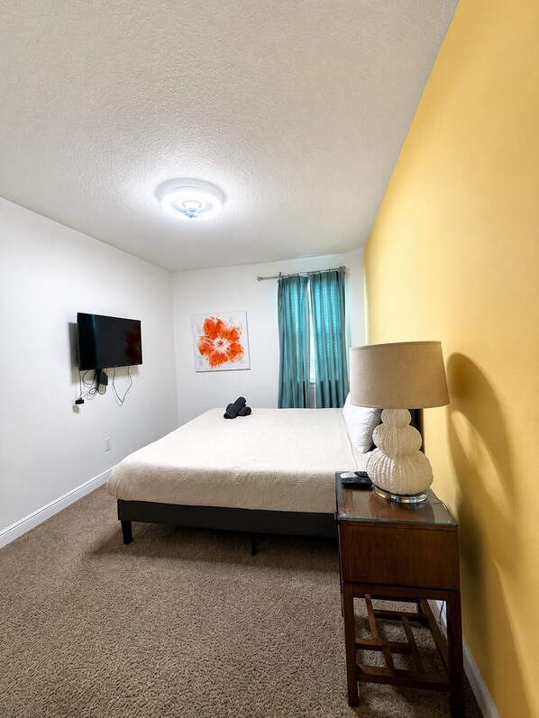 A hotel room with a bed, a flat-screen TV mounted on the wall, a bedside table with a lamp, and a window with curtains.