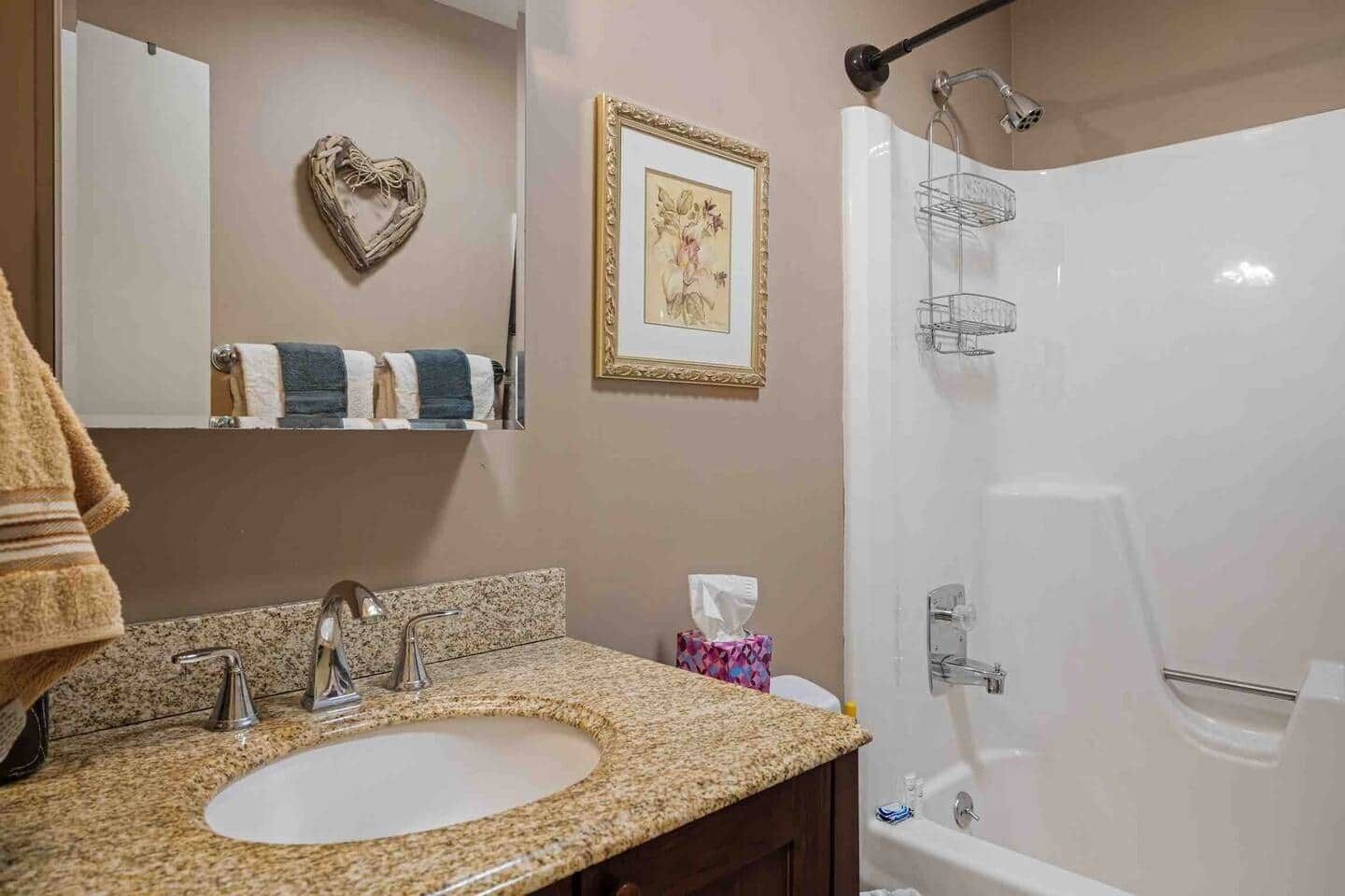 A bathroom with a granite countertop sink, a mirror, a framed picture, a towel rack with towels, and a shower with a glass enclosure.