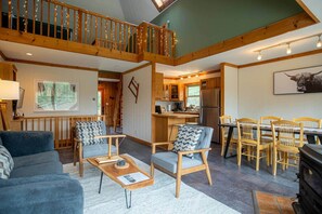 A spacious living area with a high ceiling, wooden beams, a dining table with chairs, a kitchen with modern appliances, and a cozy seating area with a sofa and armchairs.