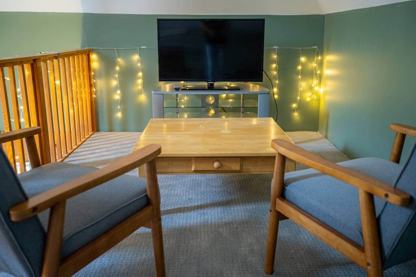 Ein gemütliches Zimmer mit Holztisch, zwei blau gepolsterten Stühlen, einem Flachbildfernseher und Lichterkette.