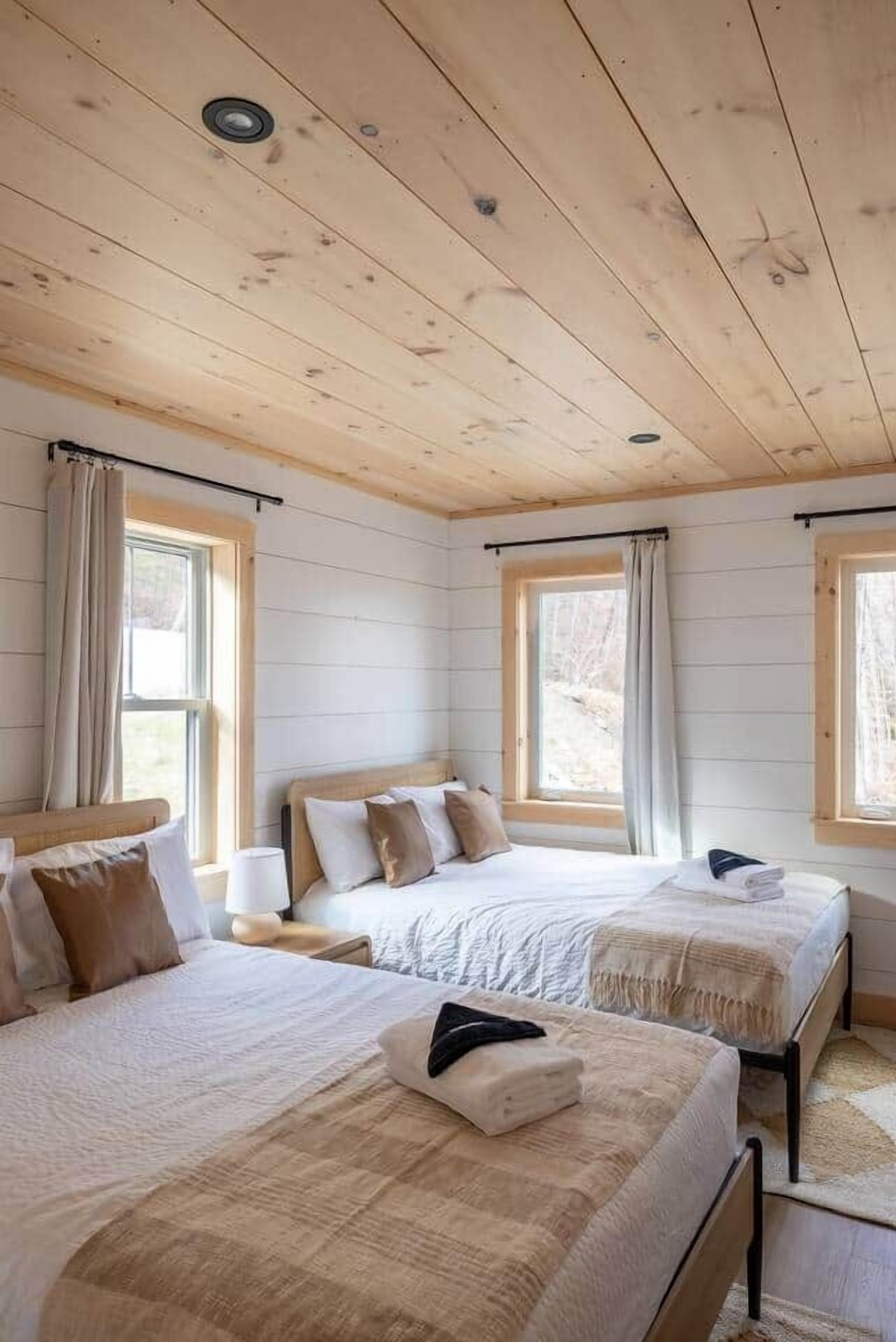 Une chambre avec deux lits, un plafond en bois et de grandes fenêtres avec des rideaux.