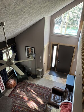 Ein Zimmer mit hohen Decken, einem Deckenventilator, einem braunen Ledersofa, einem gemusterten Teppich und einem großen Fenster mit Blick auf Bäume.