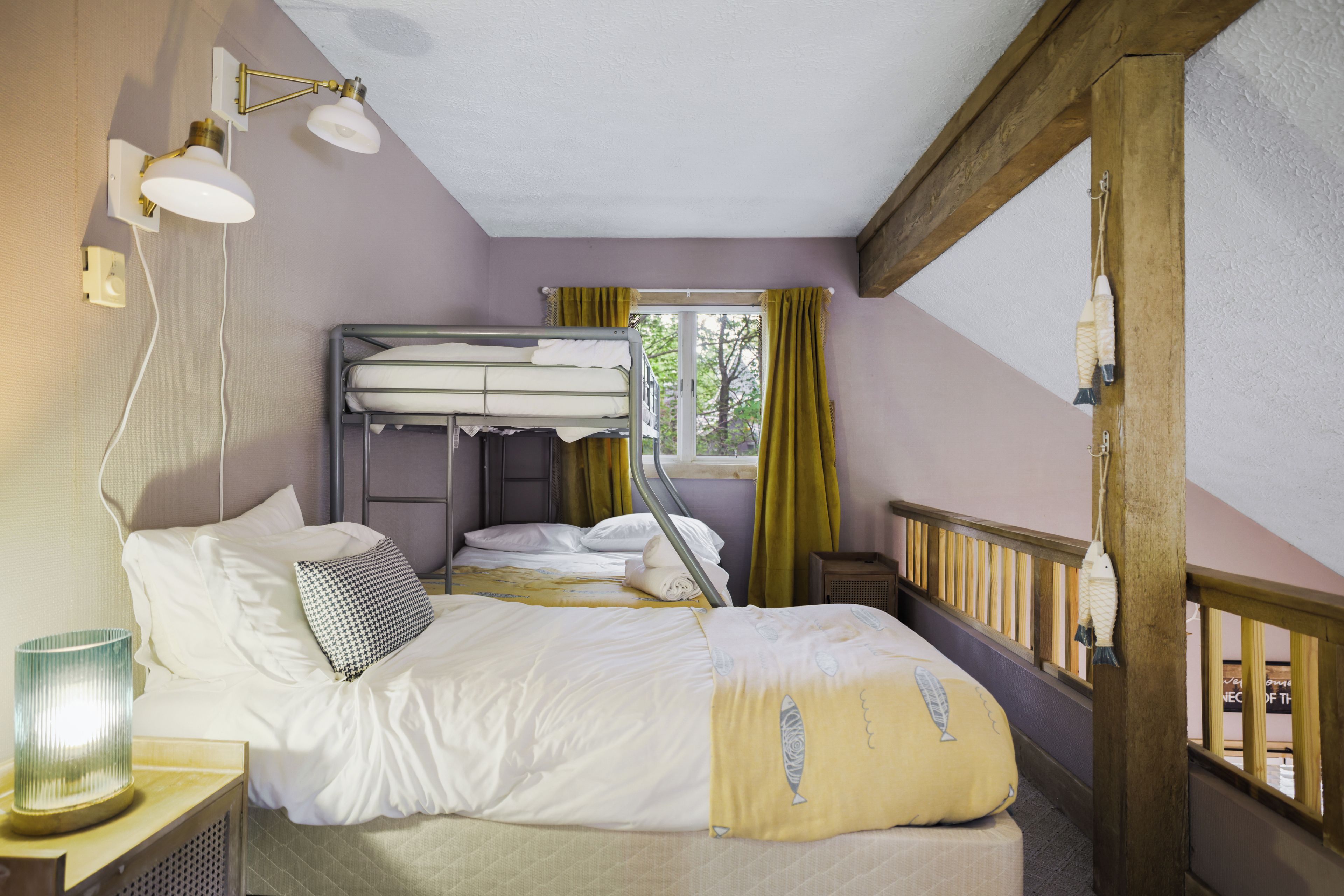Een kamer met stapelbedden, een nachtkastje met lamp, een raam met gordijnen en uitzicht op bomen.