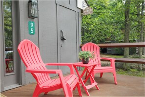 Eine Holzterrasse mit zwei roten Adirondack-Stühlen und einem kleinen Tisch mit einem Blumentopf. Ein Schild mit der Aufschrift 'The Porch Sit & Relax' ist über der Tür sichtbar.
