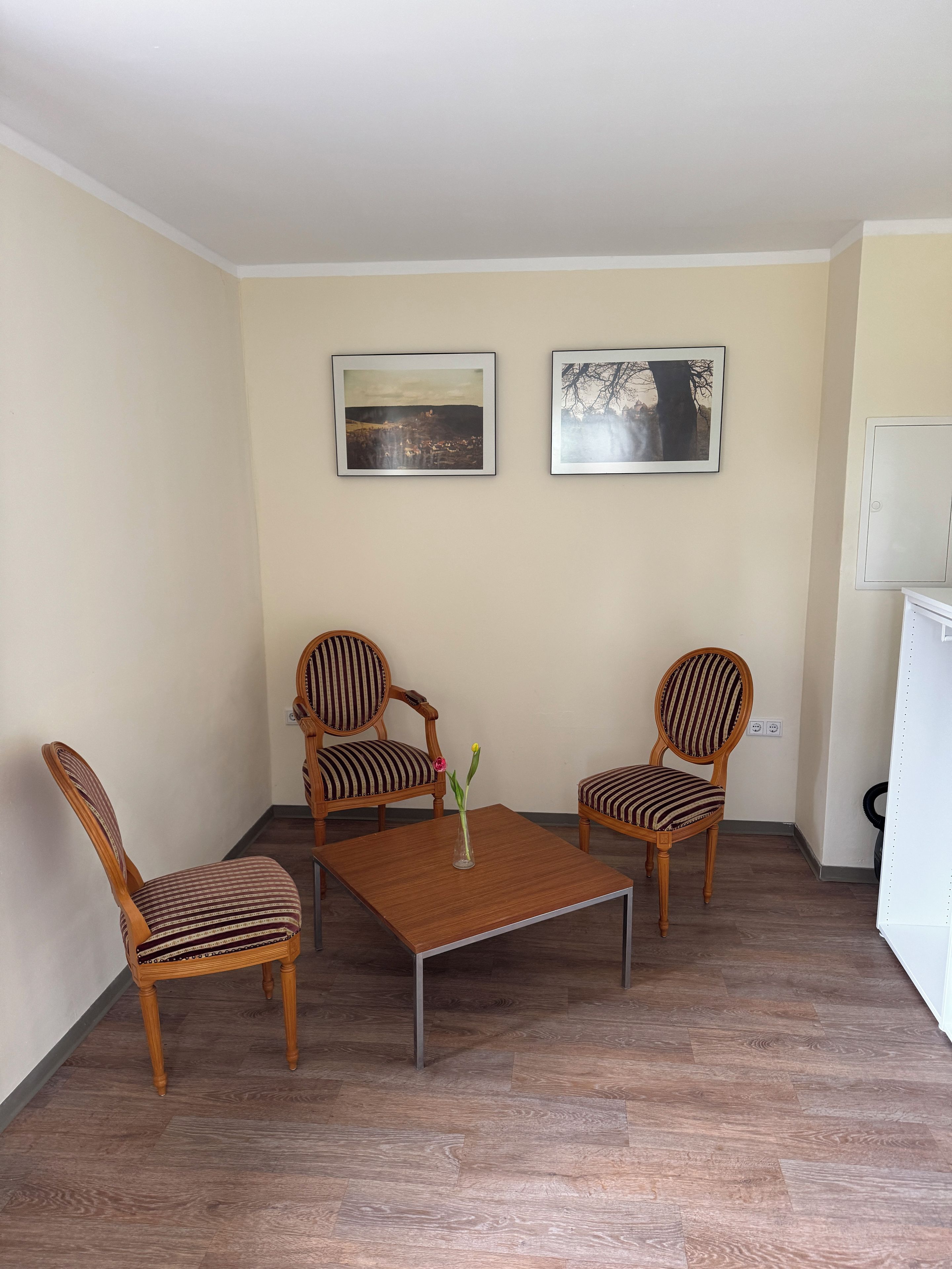 A room with two wooden chairs with striped upholstery, a square wooden table, and two framed pictures on the wall.
