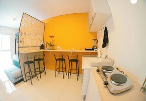 A compact kitchen with a bar area, a sink, a blender, and a coffee maker. There are three stools and a potted plant.
