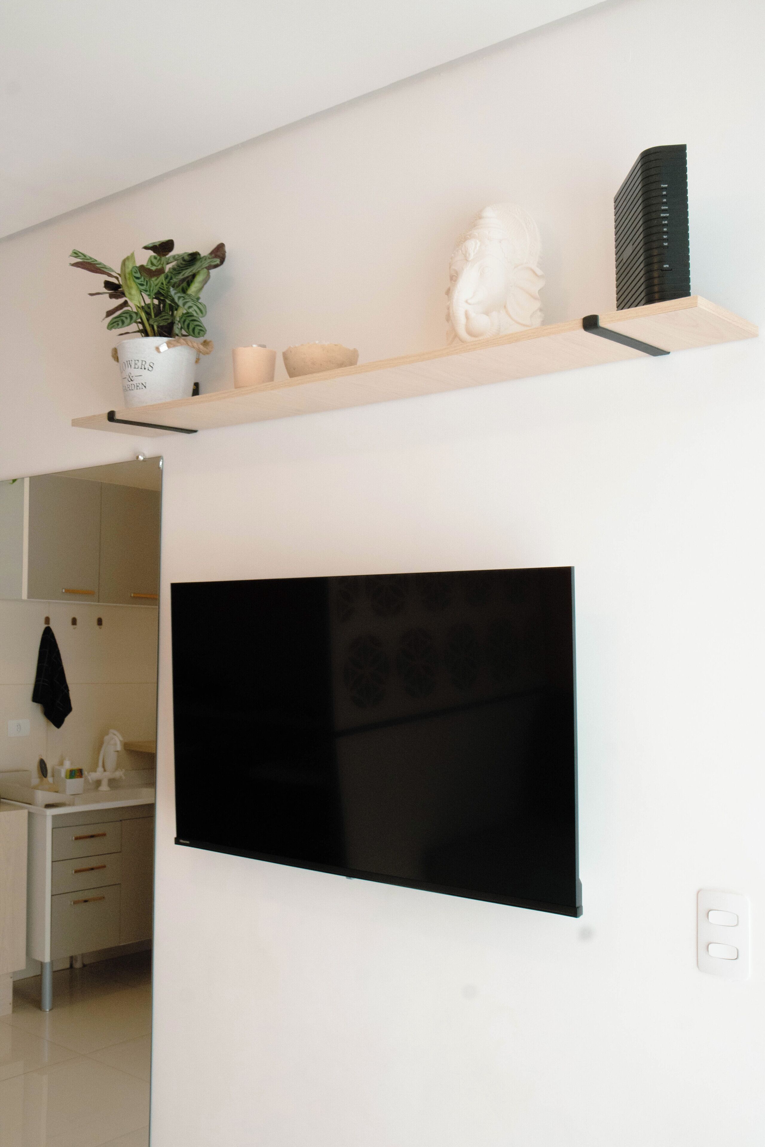 Uma cozinha moderna com uma televisão de tela plana na parede, uma prateleira de madeira acima com objetos decorativos e um vaso de planta.