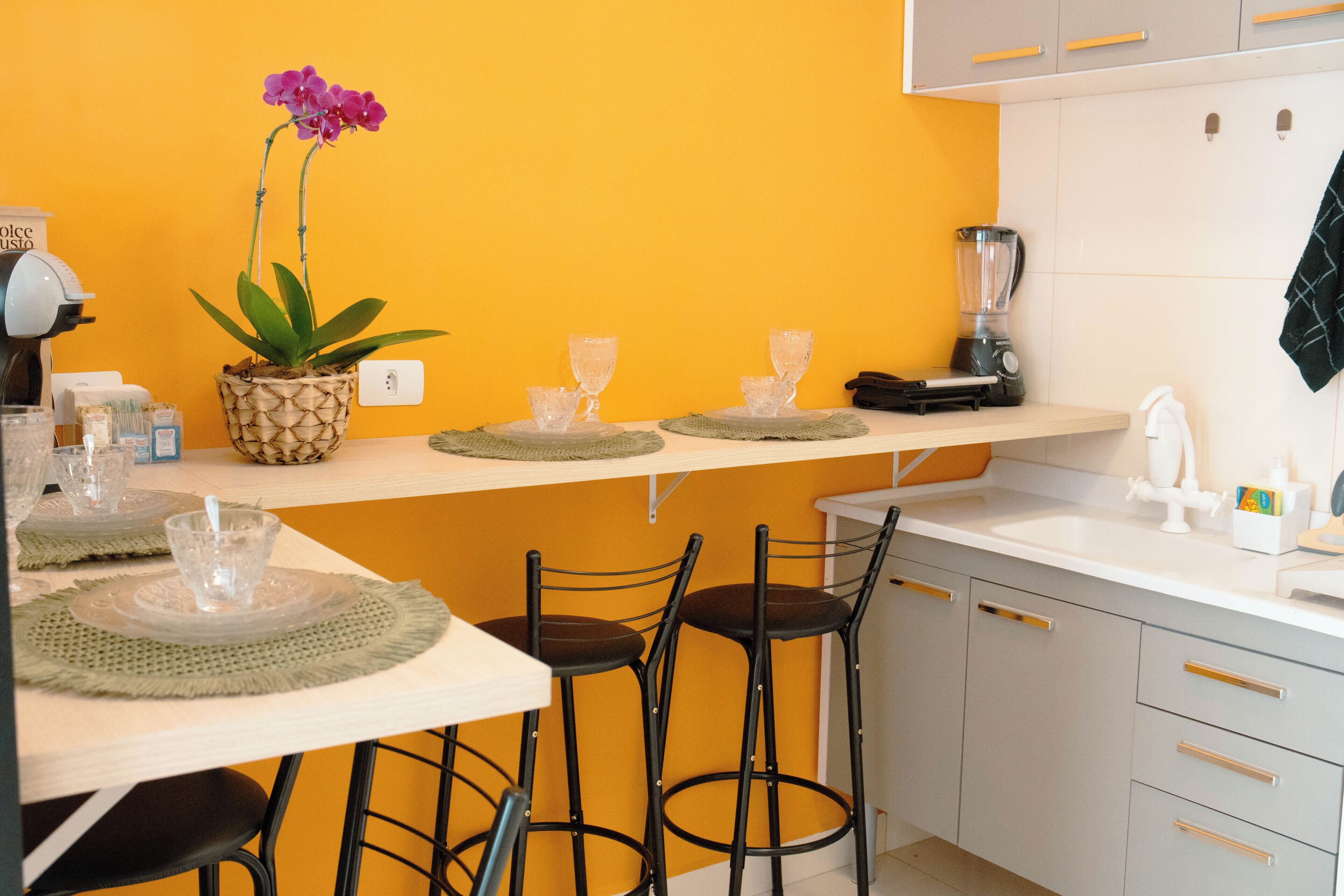 Cozinha com parede amarela, área de bar com banquetas, liquidificador e uma orquídea em vaso.