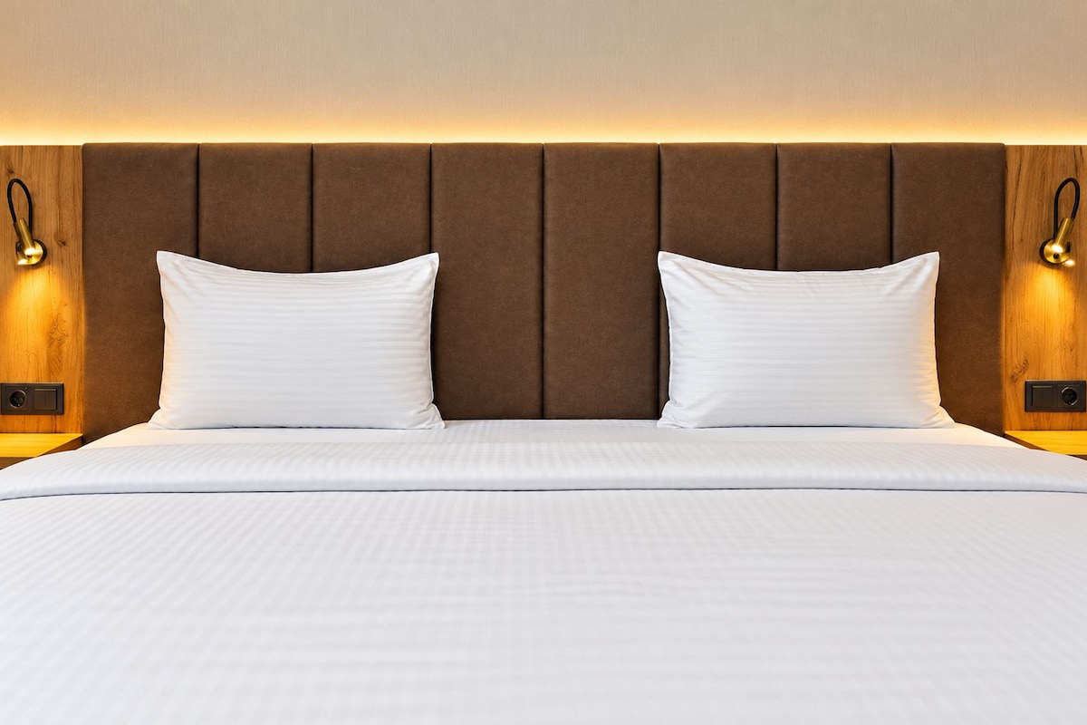 A hotel room with a large bed, two pillows, a wooden headboard, and wall-mounted lamps.