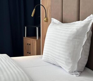 Una cama con un almohadón blanco y una manta a rayas, una mesita de noche con una lámpara, y un armario de madera al fondo.