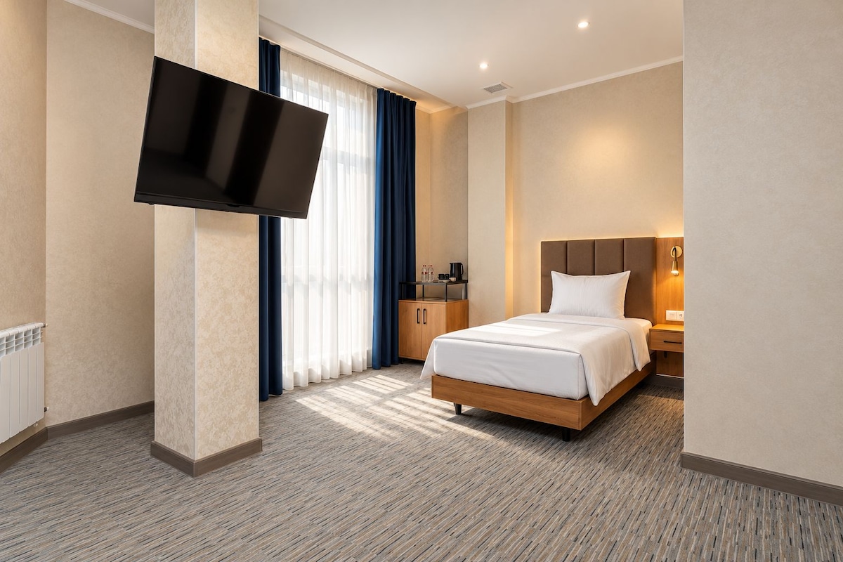 A hotel room with a bed, a flat-screen TV mounted on the wall, a wooden nightstand, a bedside table with a lamp, and a window with curtains.