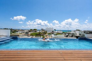 A swimming pool with a person swimming, surrounded by a wooden deck and overlooking a coastal area with buildings. - Modern Apartment Pool & Balcony (Playa del Carmen)