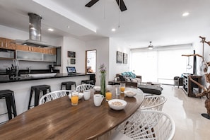 A modern kitchen with a dining area, featuring a wooden table, bar stools, and a seating area with a sofa and coffee table. - Modern Apartment Pool & Balcony (Playa del Carmen)