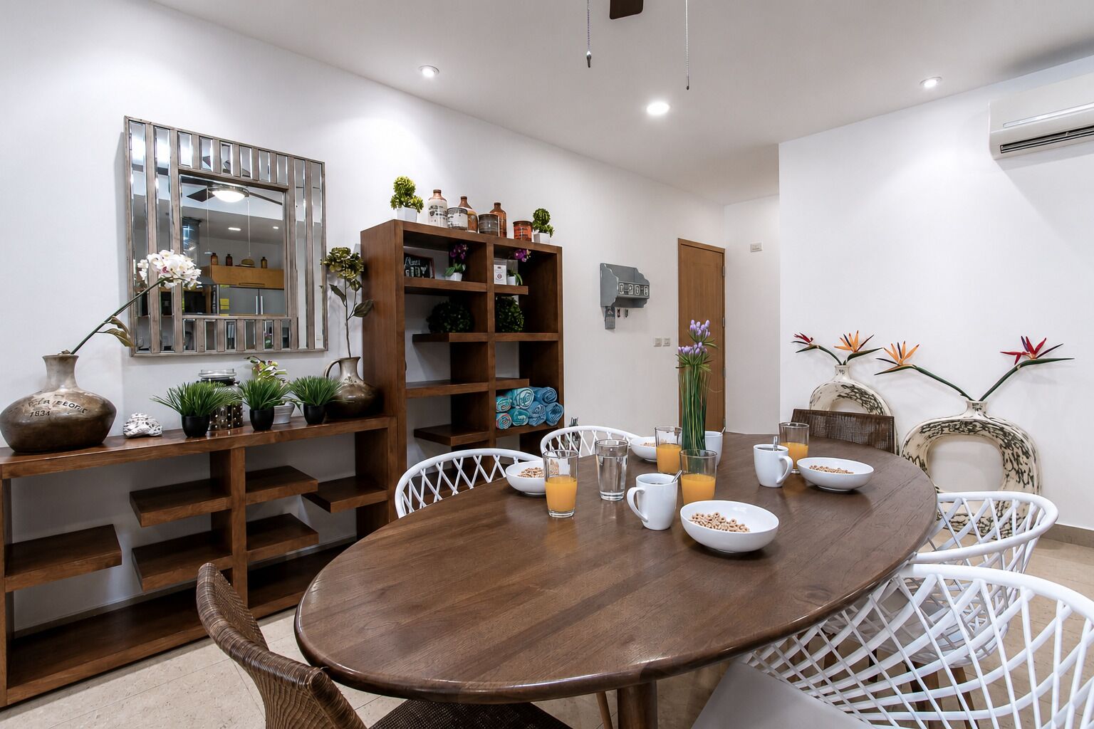 Un comedor con una mesa redonda de madera, sillas blancas y una estantería con varios objetos.
