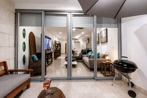 A modern living room with a glass door leading to a dining area, a sofa, a coffee table with a glass of wine, and a barbecue grill. - Modern Apartment Pool & Balcony (Playa del Carmen)