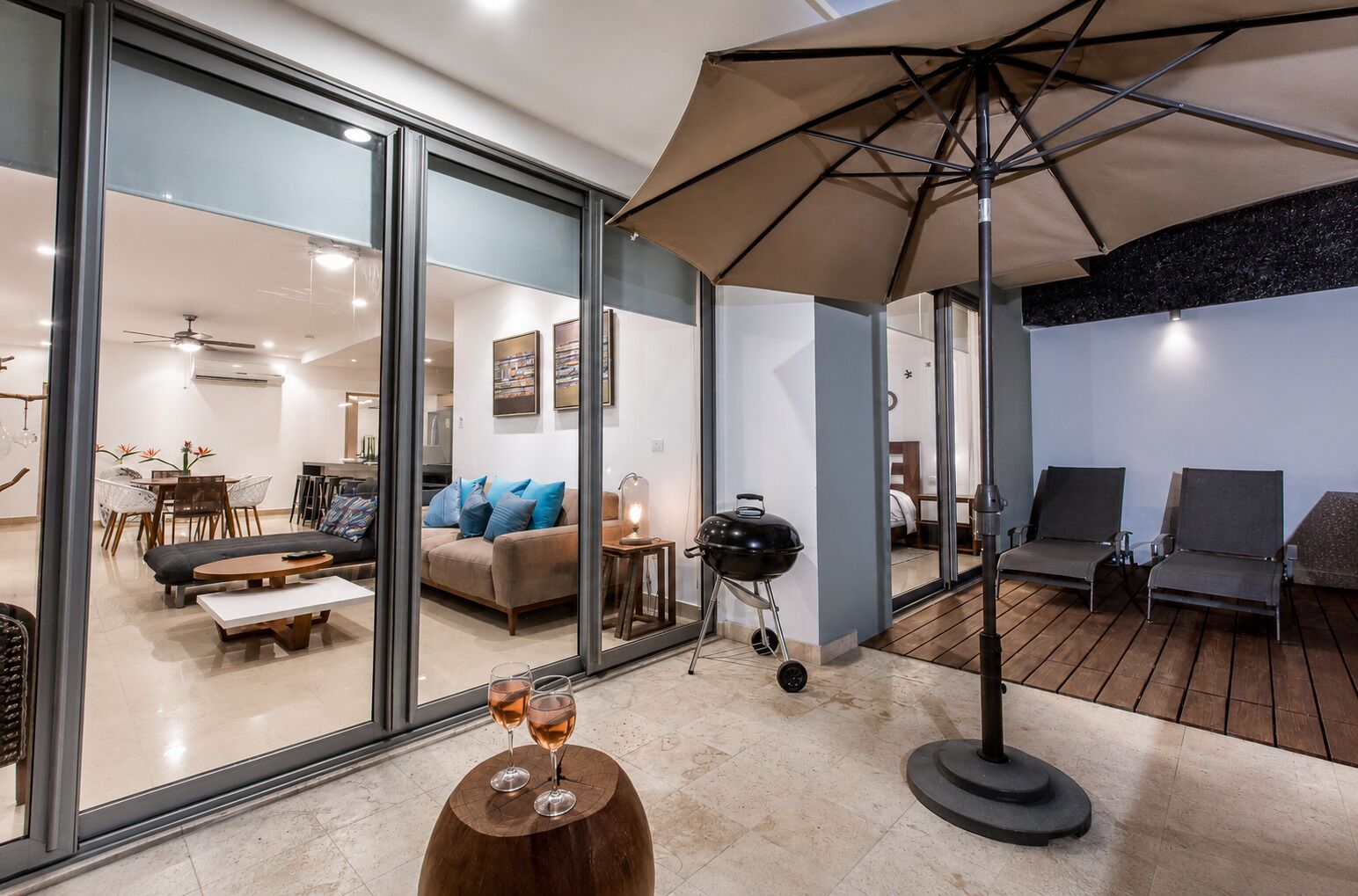 Una sala de estar moderna con pared de vidrio, una mesa de madera con vasos, una parrilla y un área de patio con sillas.