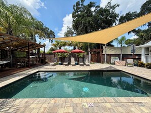 Une piscine entourée d’une terrasse en bois et d’un espace de repos couvert, avec des chaises longues et des parasols.