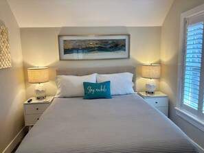 Un lit bien fait, recouvert d’une couette blanche et agrémenté de deux oreillers, dont l’un porte un motif imprimé. On y voit deux tables de chevet avec des lampes, une œuvre d’art encadrée au-dessus du lit, et une fenêtre avec des stores sur le côté droit.