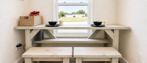 Une table-banc en bois avec deux bols et deux tasses, un panier et une fenêtre donnant sur un bâtiment et des arbres.