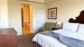 A hotel room with a bed, a chair, a nightstand, a lamp, a painting, and a bathroom visible through an open door. - Walking distance from the World Famous, Indian Wells Tennis Gardens (La Quinta)