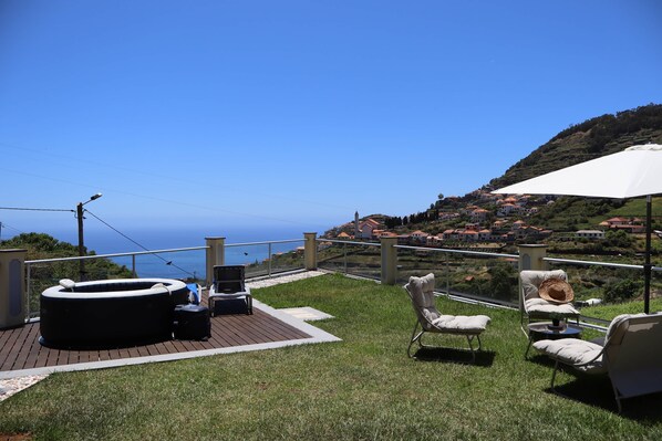 A deck with outdoor furniture, a hot tub, and a view of a coastal town. - Sunset Villa Madeira (Ribeira Brava)