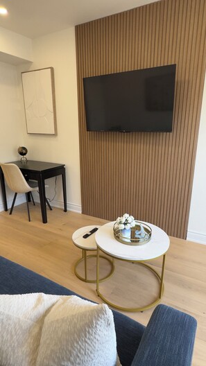 Ein modernes Wohnzimmer mit Flachbildfernseher an einer Wand aus Holzpaneelen, einem runden Couchtisch mit Tablett, einer Couch und einem Essbereich mit einem Stuhl und einem kleinen Tisch.