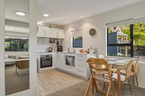 A modern kitchen with white cabinets, stainless steel appliances, and a dining area with wooden chairs and a glass table. - Amalia Cottage - Lake Okareka, Huge Covered Deck, Sleeps 4 (Rotorua)