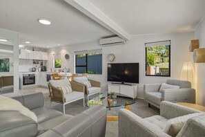 A modern living room with a flat-screen TV, grey sofas, wicker chairs, and a glass coffee table. - Amalia Cottage - Lake Okareka, Huge Covered Deck, Sleeps 4 (Rotorua)