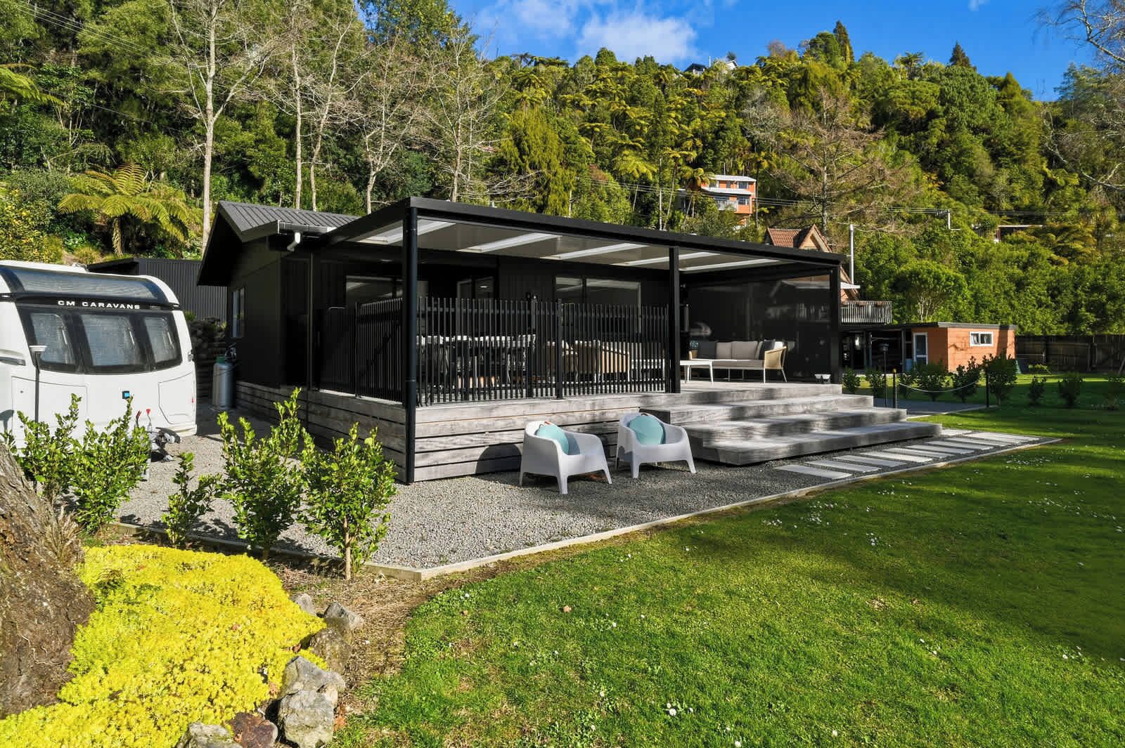 A modern outdoor seating area with a covered patio, a white caravan parked nearby, and a lush green landscape in the background.